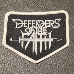 Può includere: Toppa in bianco e nero con la scritta "DEFENDERS OF THE FAITH" e un disegno di spada. La toppa ha un bordo bianco ed è su una superficie strutturata. Adatta per giacche o borse.