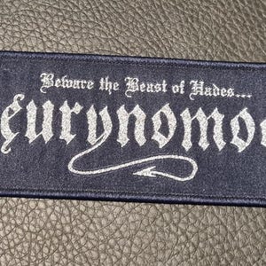 May include: A dark blue rectangular patch featuring white embroidered text and illustrations. The text reads "Beware the Beast of Hades... Eurynomos." Two devil figures with pitchforks and flames are on either side. Likely for clothing or accessories.