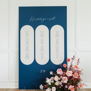 May include: A navy blue wedding seating chart with three white arched panels listing guest names. The text "We saved you a seat!" is written in cursive at the top. The initials "J|S" are at the bottom. A floral arrangement is in the lower right.
