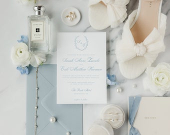 Puede incluir: Un conjunto de invitación de boda con un sobre azul claro, una tarjeta de invitación blanca con una corona azul y el texto "Sarah Anne Zuccolo y Erik Matthew Kroman", una caja blanca con un anillo dentro y un libro blanco con el texto "Her Vows".