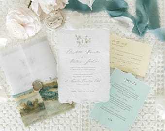 Op de afbeelding: Trouwuitnodigingsset met een hoofduitnodiging met bloemenwapen, een RSVP-kaart, een detailkaart en een landschapsbedrukte envelop. De set bevat een ringdoosje en is omgeven door witte bloemen en blauwe linten.