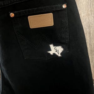 May include: Black denim jeans with a brown leather Wrangler patch on the back pocket and a white Texas-shaped patch with the letters "SV" inside.