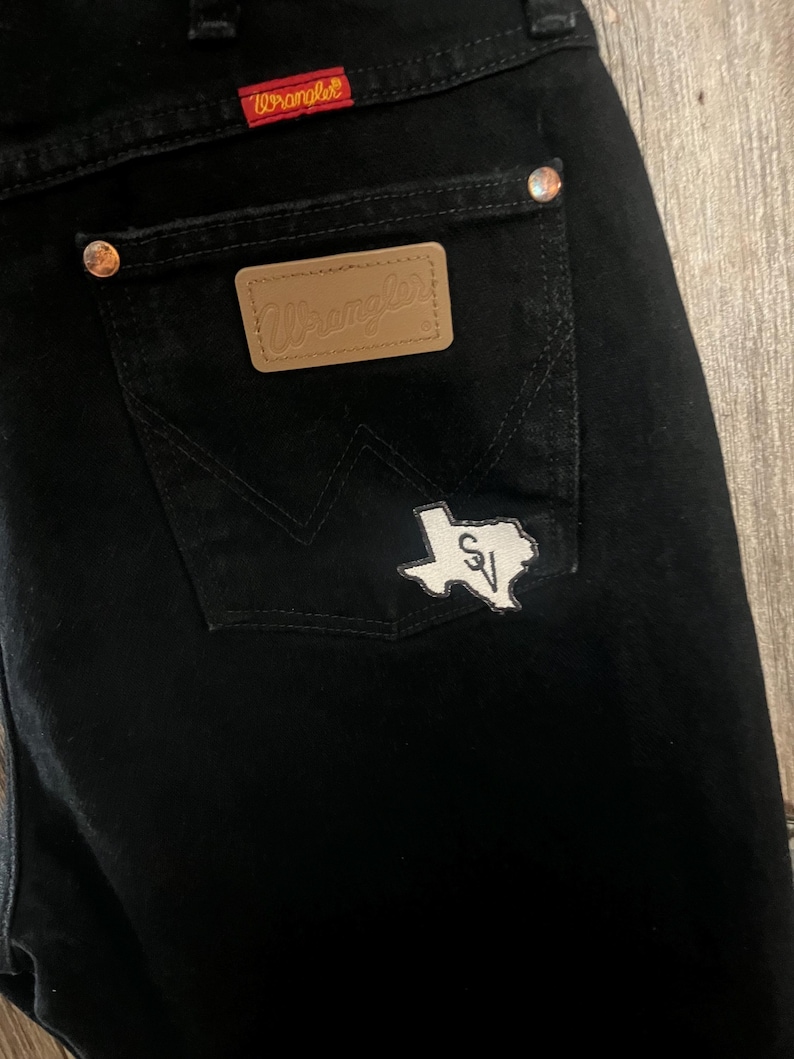 May include: Black denim jeans with a brown leather Wrangler patch on the back pocket and a white Texas-shaped patch with the letters "SV" inside.