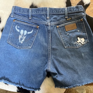May include: Blue denim Wrangler shorts with a raw hem. The left back pocket features a white bull skull design, while the right pocket has a Texas-shaped patch. The shorts have a classic five-pocket design.