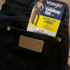 May include: Black denim jeans with a brown leather Wrangler label and a yellow and white tag that reads "The Original Cowboy Cut Jean" and "Natural Rise 14MWZ Slim Fit Tapered Leg Fits Over Boots".