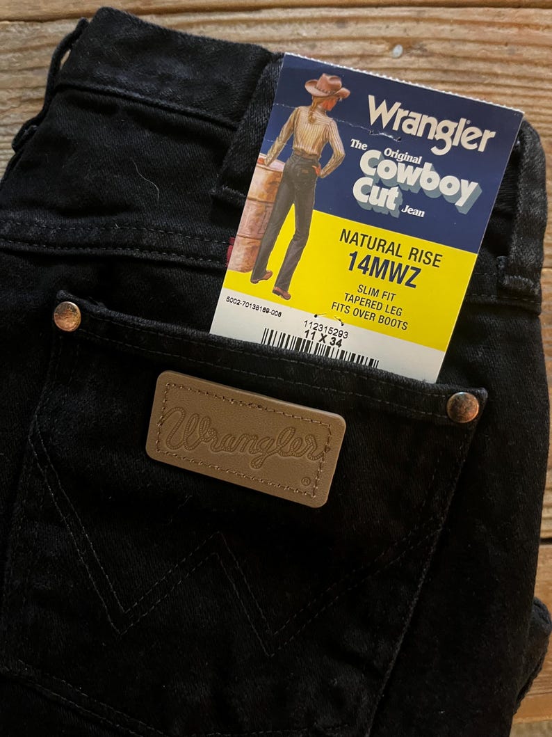 May include: Black denim jeans with a brown leather Wrangler label and a yellow and white tag that reads "The Original Cowboy Cut Jean" and "Natural Rise 14MWZ Slim Fit Tapered Leg Fits Over Boots".