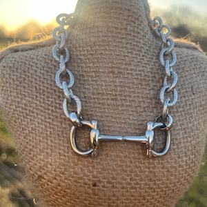 May include: Silver-toned necklace featuring a horse bit design. The necklace has a thick chain with textured links and a classic horse bit pendant. The jewelry is displayed on a burlap-covered bust, with a blurred outdoor background.