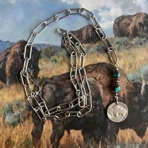 May include: Silver-toned chain necklace with a pendant featuring a buffalo nickel. The necklace has turquoise and dark brown beads. The background shows a painting of bison in a grassy field.