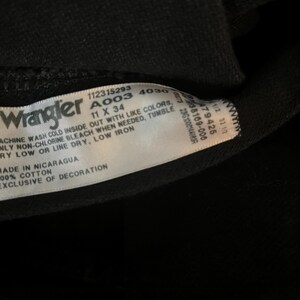 May include: A black Wrangler jeans care label with white text. The label includes the size 11 x 34, the style number A003 4030, and washing instructions. The label also states "Made in Nicaragua" and "100% Cotton, Exclusive of Decoration."