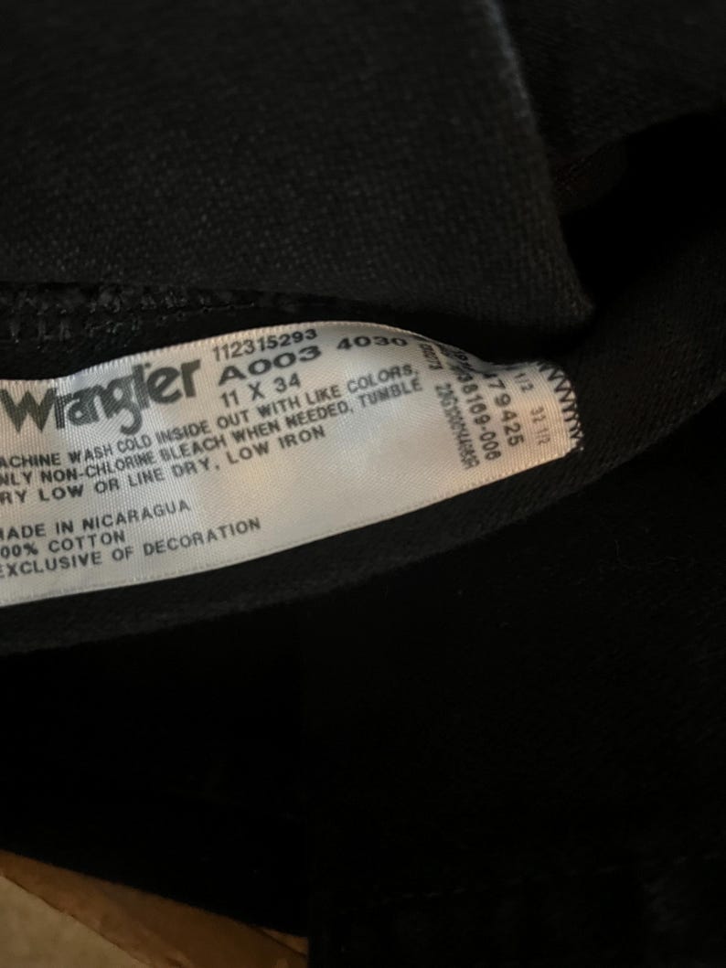 May include: A black Wrangler jeans care label with white text. The label includes the size 11 x 34, the style number A003 4030, and washing instructions. The label also states "Made in Nicaragua" and "100% Cotton, Exclusive of Decoration."