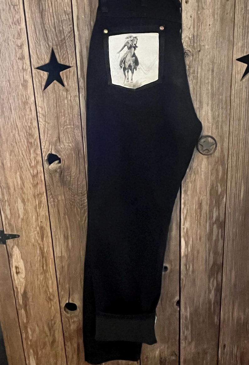 May include: Black denim jeans with a white patch pocket featuring a black and white image of a person riding a horse.