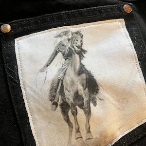 May include: A pair of black jeans with a white patch on the back pocket. The patch features a black and white image of a cowboy riding a horse.