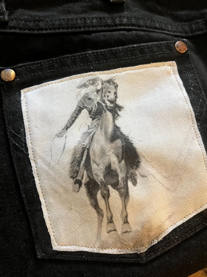 May include: A pair of black jeans with a white patch on the back pocket. The patch features a black and white image of a cowboy riding a horse.
