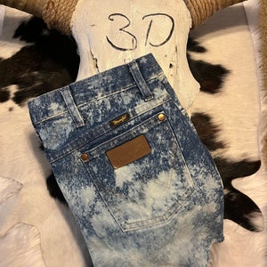 May include: Blue denim Wrangler shorts with a bleached, faded design. The shorts have a brown leather Wrangler patch on the back pocket and gold-colored rivets. The shorts are displayed on a cowhide rug with a skull in the background.