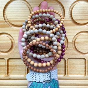 May include: A hand holding a stack of wooden bead bracelets in various colours, including brown, beige, and purple. The bracelets are stacked on top of each other and are arranged in a circular pattern.