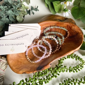 May include: A wooden bowl filled with five handmade gemstone bracelets. The bracelets are made with various stones, including green aventurine, amethyst, rose quartz, and wood beads. The bracelets are stacked on top of each other in the bowl. The bowl is sitting on a wooden surface. The text on the tag reads "Sunshine + Ivy Handmade Bead Jewelry".