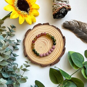 May include: A multi-coloured gemstone bracelet with pink lava beads. The bracelet is on a wooden surface with green leaves in the background.