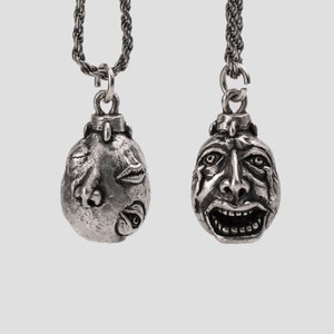 Puede incluir: Dos colgantes de plata en cadenas, cada uno representando una cara diferente. Un colgante presenta una cara con nariz, boca y ojos, mientras que el otro colgante presenta una cara con la boca abierta de par en par y las cejas fruncidas.