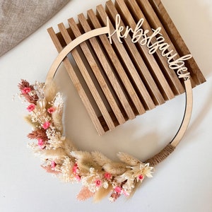 Könnte beinhalten: Ein Holzkranz mit getrockneten Blumen in Rosa, Creme und Beige. Der Kranz hat ein Holzschild mit der Aufschrift "Herbstzauber". Der Kranz ist auf einem hölzernen Lattenhintergrund angebracht.