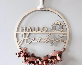 May include: A circular wooden wall hanging featuring the text "HALLO" and a pumpkin outline on the top half, with the word "Herbst" in script below. The bottom arc is decorated with dried pink and dark red flowers, suspended by a white ribbon.