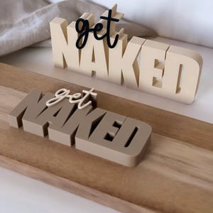 Könnte beinhalten: Zwei dekorative Schilder mit den Worten "get NAKED". Das obere Schild ist beige mit schwarzem Schreibschrifttext, und das untere Schild ist braun mit cremefarbenem Schreibschrifttext. Beide Schilder befinden sich auf einem Holztablett.