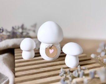 May include: Three smooth white mushroom-shaped figurines. The central, larger mushroom features a small, round, brown paper tag tied with a thin white string, displaying a single light pink heart printed in the center of the tag.