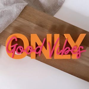 Pode incluir: Letreiro laranja "ONLY" com letras rosa "Good Vibes". O letreiro est&aacute; colocado sobre uma t&aacute;bua de madeira, com um tecido de cor clara ao fundo. As letras s&atilde;o grossas e se destacam do fundo.