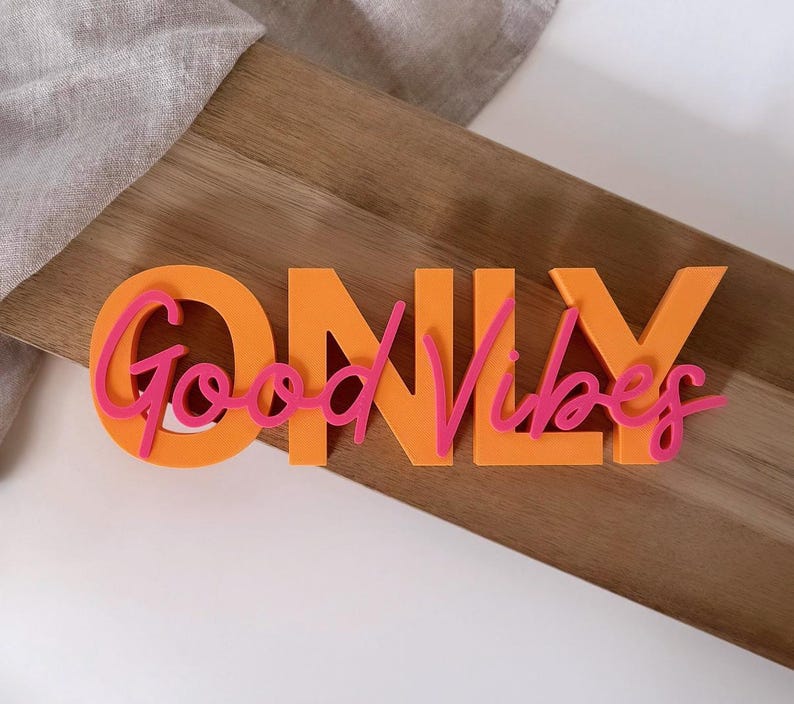 Pode incluir: Letreiro laranja "ONLY" com letras rosa "Good Vibes". O letreiro est&aacute; colocado sobre uma t&aacute;bua de madeira, com um tecido de cor clara ao fundo. As letras s&atilde;o grossas e se destacam do fundo.