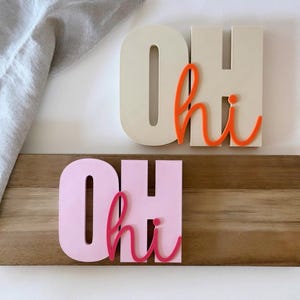 Könnte beinhalten: Zwei Holzschilder mit den Worten "OH hi" in einer verspielten Schriftart. Ein Schild ist cremefarben mit orangefarbenen Buchstaben, und das andere ist rosa mit pinkfarbenen Buchstaben. Die Schilder werden auf einem Holzbrett angezeigt.