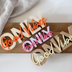 Pode incluir: Recortes de madeira que dizem "Good Vibes Only" em v&aacute;rias cores. As palavras est&atilde;o em uma mistura de fontes e cores, incluindo laranja, rosa e amarelo. Os recortes s&atilde;o dispostos em uma superf&iacute;cie de madeira.