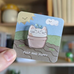 May include: A clear acrylic pin featuring a smiling cat in a flower pot with a sprout. The pin is set against a blue sky background with a sun and clouds. The text "PULP AND POPPY" is printed at the bottom.