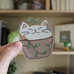 May include: A clear sticker with a cartoon illustration of a cat with orange and white fur, smiling, inside a brown pot with green vines. The cat has a green sprout growing from its head.