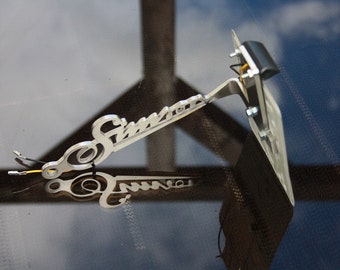 May include: A silver motorcycle number plate holder with the word "Simota" engraved on it. The holder has a black light attached to it.