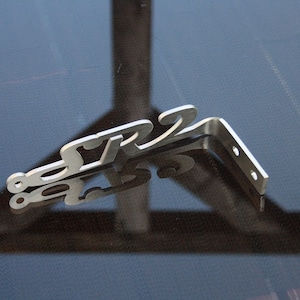 Puede incluir: Un soporte de metal plateado con la palabra "SR2" recortada en el metal. El soporte está diseñado para ser montado en una pared u otra superficie.