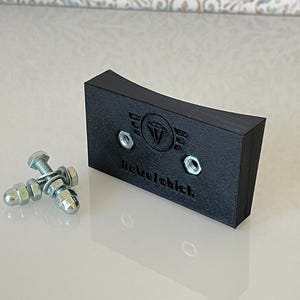 Könnte beinhalten: Ein schwarzes, rechteckiges Objekt mit abgerundeten Kanten, mit dem Text "nickelchick" und einem Logo mit Diamant und Flügeln. Zwei Muttern und Schrauben befinden sich neben dem Objekt. Der Hintergrund ist ein Blumenmuster.