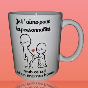 Peut inclure: Mug blanc en céramique avec un dessin noir et rouge de deux personnages et du texte en français. La tasse a une anse incurvée et une finition brillante. Le texte se lit "Jet' aime pour ta personnalité mais ce cul est un énorme bonus."