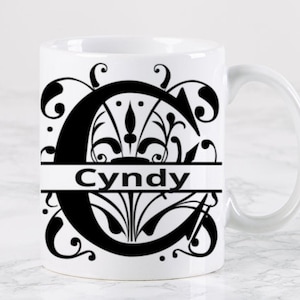 Peut inclure: Une tasse en céramique blanche avec un motif noir et blanc représentant une lettre C stylisée avec des accents floraux et le nom "Cindy" imprimé en dessous.