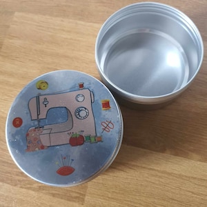 Könnte beinhalten: Eine runde Metalldose mit einem blauen und weißen Design, das eine Nähmaschine, Garnrollen und Nadeln zeigt. Der Deckel der Dose ist mit einer Nähmaschine verziert, der Boden der Dose ist schlicht.