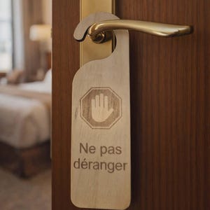 Può includere: Un cartello di legno per la porta con la scritta francese "Ne pas déranger". Il cartello presenta un simbolo di mano all'interno di un segnale di stop. La maniglia della porta è dorata ed è attaccata a una porta di legno marrone.