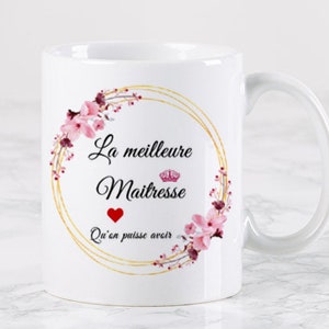 Könnte beinhalten: Weiße Keramiktasse mit einem goldenen Blumengirlanden-Design. Der Text "La meilleure Maitresse" ist in Schwarz mit einem roten Herzen und einer Krone über dem Wort "Maitresse". Der Text "Qu'en puisse avoir" befindet sich unter dem Kranz.
