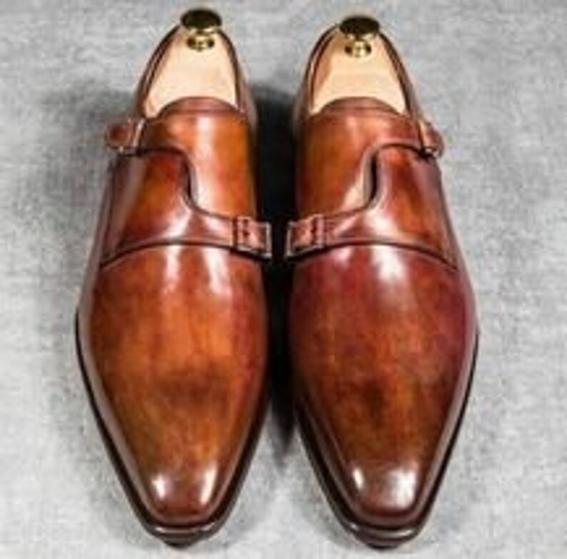 May include: A pair of brown leather monk strap shoes with a pointed toe and a polished finish.