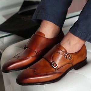 Pu&ograve; includere: Un paio di scarpe da monaco in pelle marrone con design a punta alare. Le scarpe hanno due fibbie su ogni cinturino.