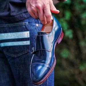 May include: A dark blue leather dress shoe with a single buckle strap. The shoe has a cap toe and a red leather sole.