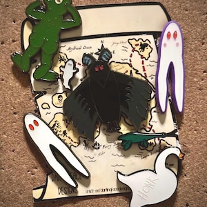 May include: Enamel pins featuring a green frog, a black moth, a white ghost, a white fork-shaped ghost, a green sea creature, and a white goose with the word "HONK". The pins are on a map-themed background.