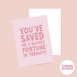 May include: A light pink greeting card featuring bold, dark pink text which reads, "YOU'VE SAVED ME A BLOODY FORTUNE IN THERAPY!" The card is placed in front of a plain white envelope, with a small circular logo in the corner reading "FOREVER BLESSED."