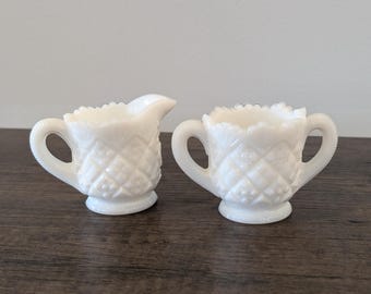 May include: A white milk glass creamer and sugar bowl set with a diamond pattern. The creamer has a handle and a spout. The sugar bowl has two handles.