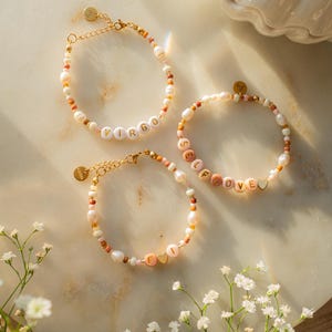 Könnte beinhalten: Drei Perlenarmbänder mit goldenen Anhängern. Die Armbänder buchstabieren "Virgo", "Self Love" und "Fi" mit einem Herz-Anhänger. Die Armbänder liegen auf einer weißen Marmorfläche mit weißen Blumen.