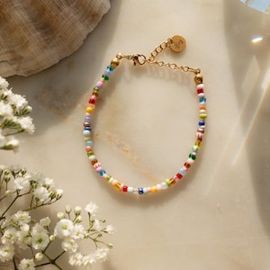 Könnte beinhalten: Ein farbenfrohes Perlenarmband mit einem goldenen Verschluss und einem kleinen goldenen Anhänger mit der Aufschrift "Freude".