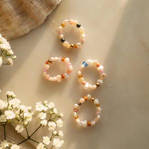 Könnte beinhalten: Vier zarte Ringe aus kleinen weißen Perlen, goldenen Perlen und bunten Glasperlen. Die Ringe sind auf einer weißen Oberfläche mit einer weißen Blume im Hintergrund angeordnet.