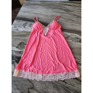 May include: A bright pink camisole-style nightgown with spaghetti straps. The bodice features a white lace trim along the neckline, and the hemline is also trimmed with white lace. The fabric appears soft and lightweight.